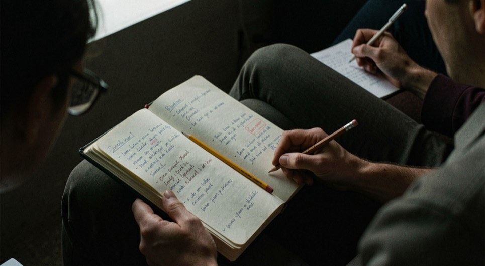 Cahier de projection annoté sur les genoux, crayon posé dans la reliure, salle sombre autour, notes prises dans la pénombre de l'écran.